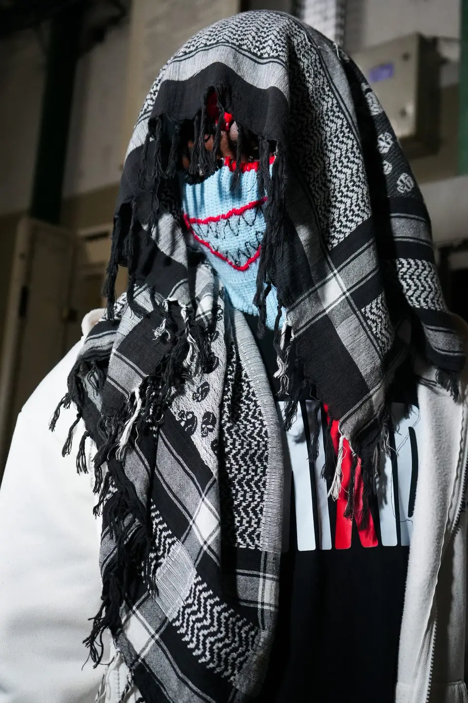 Personne portant une cagoule bleu clair à la bouche brodée de rouge, sous un keffieh noir et blanc